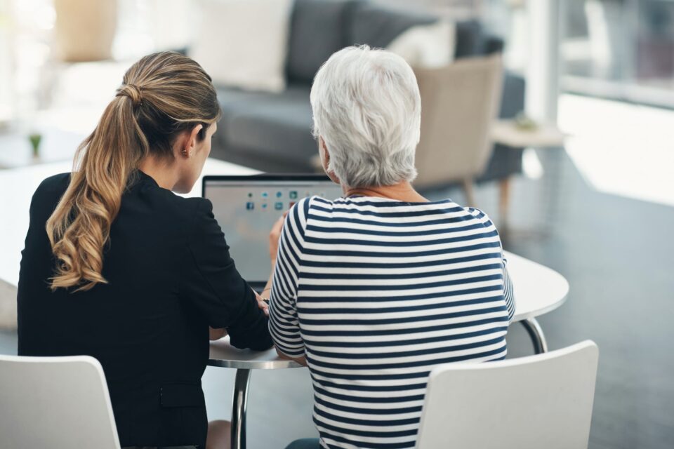 A younger woman and an older woman seated at a round table, viewed from behind, reviewing information on a laptop